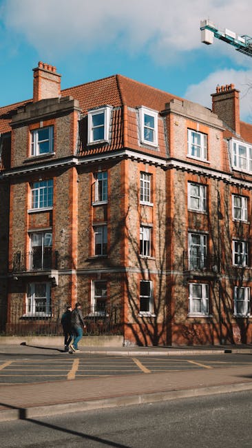 A multi-storey residential building with a traditional brick exterior, featuring white-framed sash windows and a pitched red-tiled roof, situated on a street with a concrete pavement and yellow line markings. Two individuals wearing dark clothing are walking along the sidewalk in front of the building. Shadows of leafless trees are cast across the building’s façade, indicating a sunny day with a blue sky and some cloud cover. A blue crane is visible in the upper right corner of the image, suggesting nearby construction activity. This scene captures the context of urban house removals or furniture transport, with the building presenting an ideal setting for home relocation services. Streatham Removals’ expertise in packing, loading, and transport processes for residential moves is relevant to this environment, highlighting the logistical aspects of house removals near properties like this one.